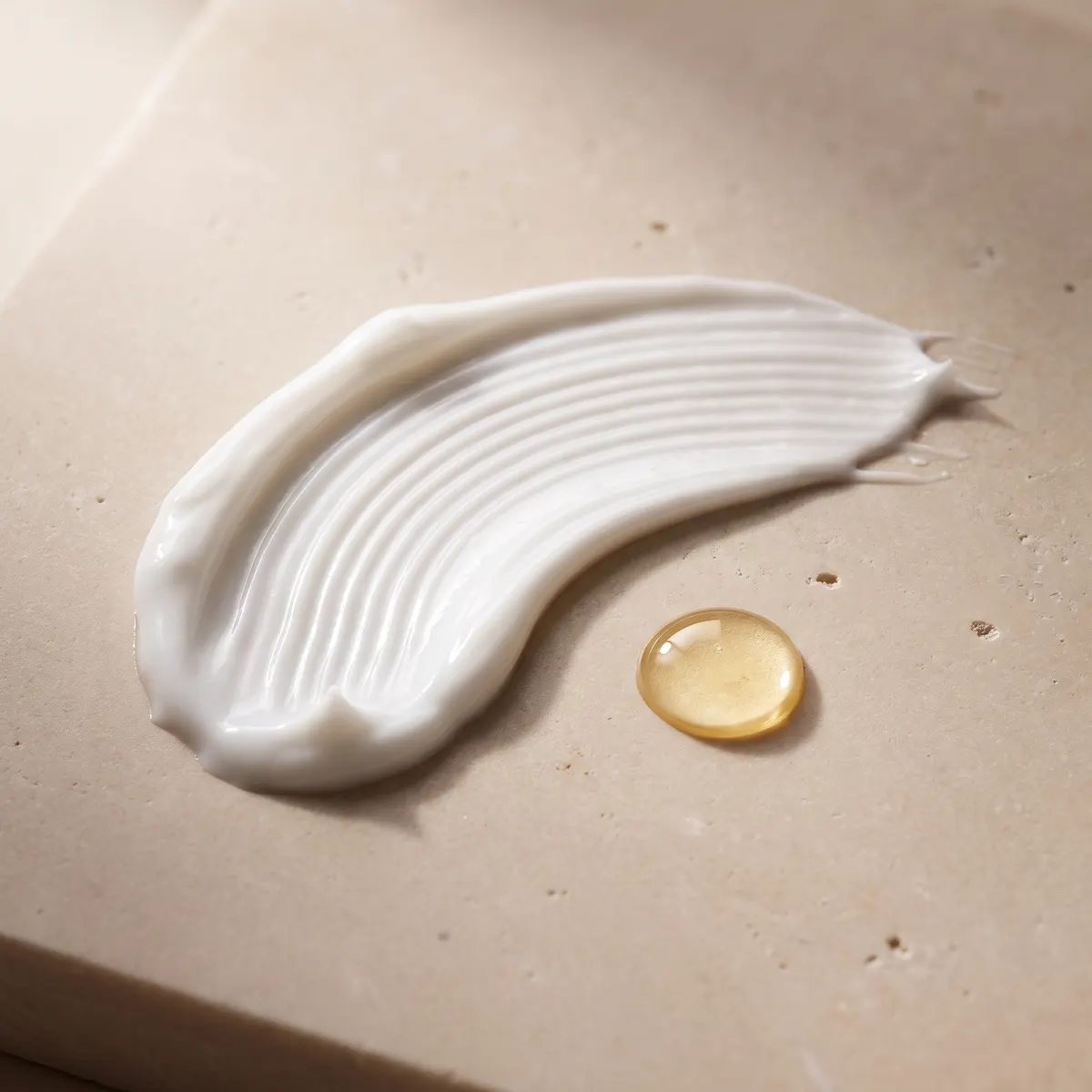 Texture macro: a swept ridge of white cream beside a golden serum drop on warm stone.